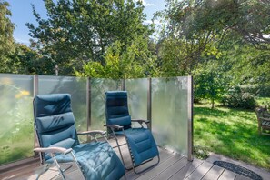 A patio area with two blue chairs and a table, surrounded by a glass fence, overlooking a lush garden. - Holiday Home Ahrenshoop with Private Terrace, Garden, and Wi-Fi (Ahrenshoop)