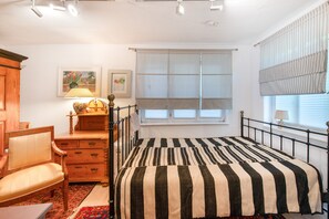 Une chambre à coucher avec un lit, une commode en bois, une chaise, une fenêtre avec des stores et un tableau accroché au mur.
