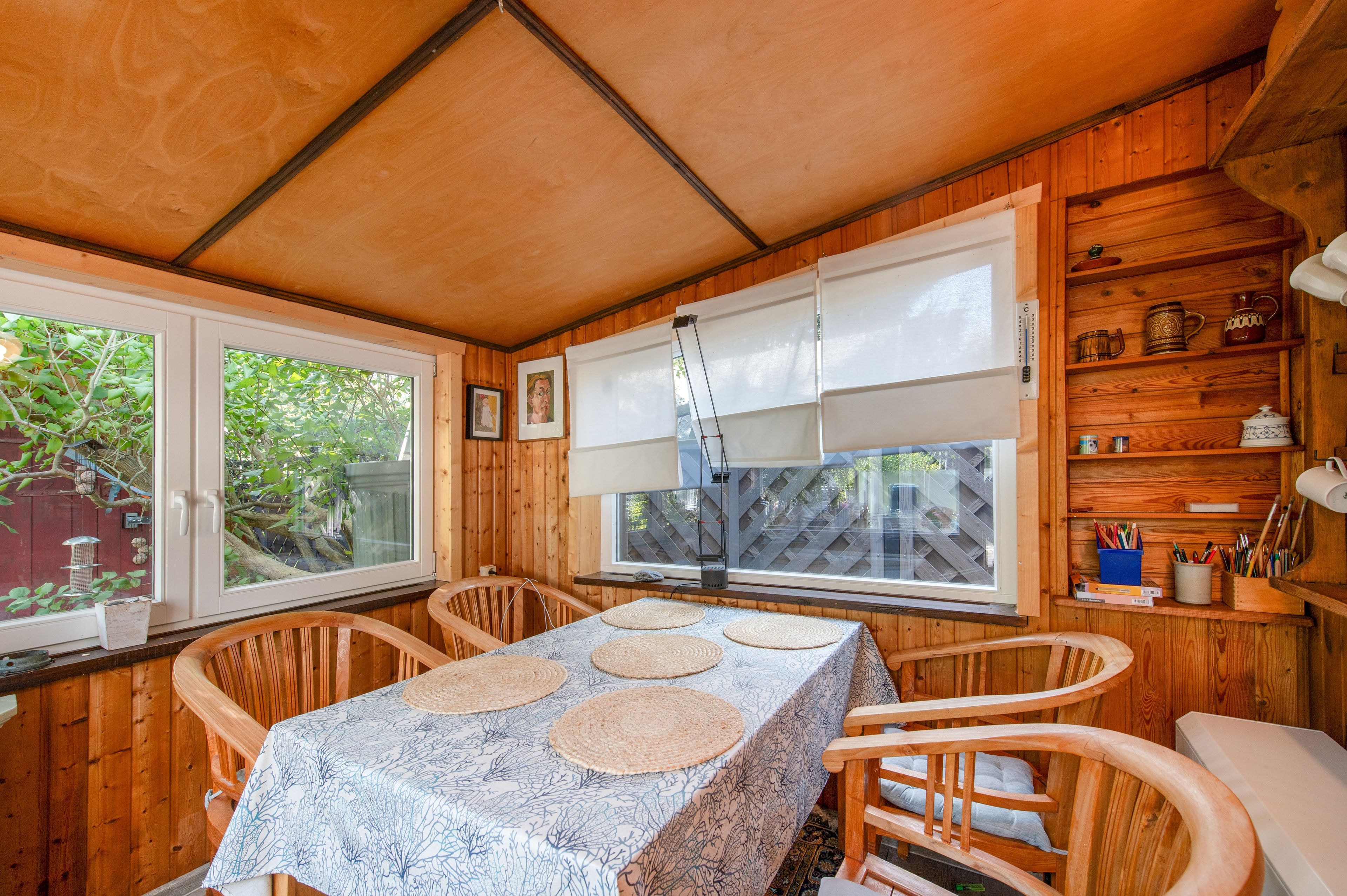 A wooden cabin interior with a dining table, chairs, a window with blinds, and a shelf with kitchenware.
