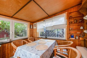A wooden cabin interior with a dining table, chairs, a window with blinds, and a shelf with kitchenware.
