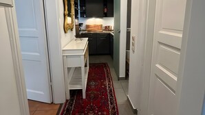 Un couloir menant à une cuisine, avec un tapis à motifs rouges, des murs blancs et un luminaire au plafond.
