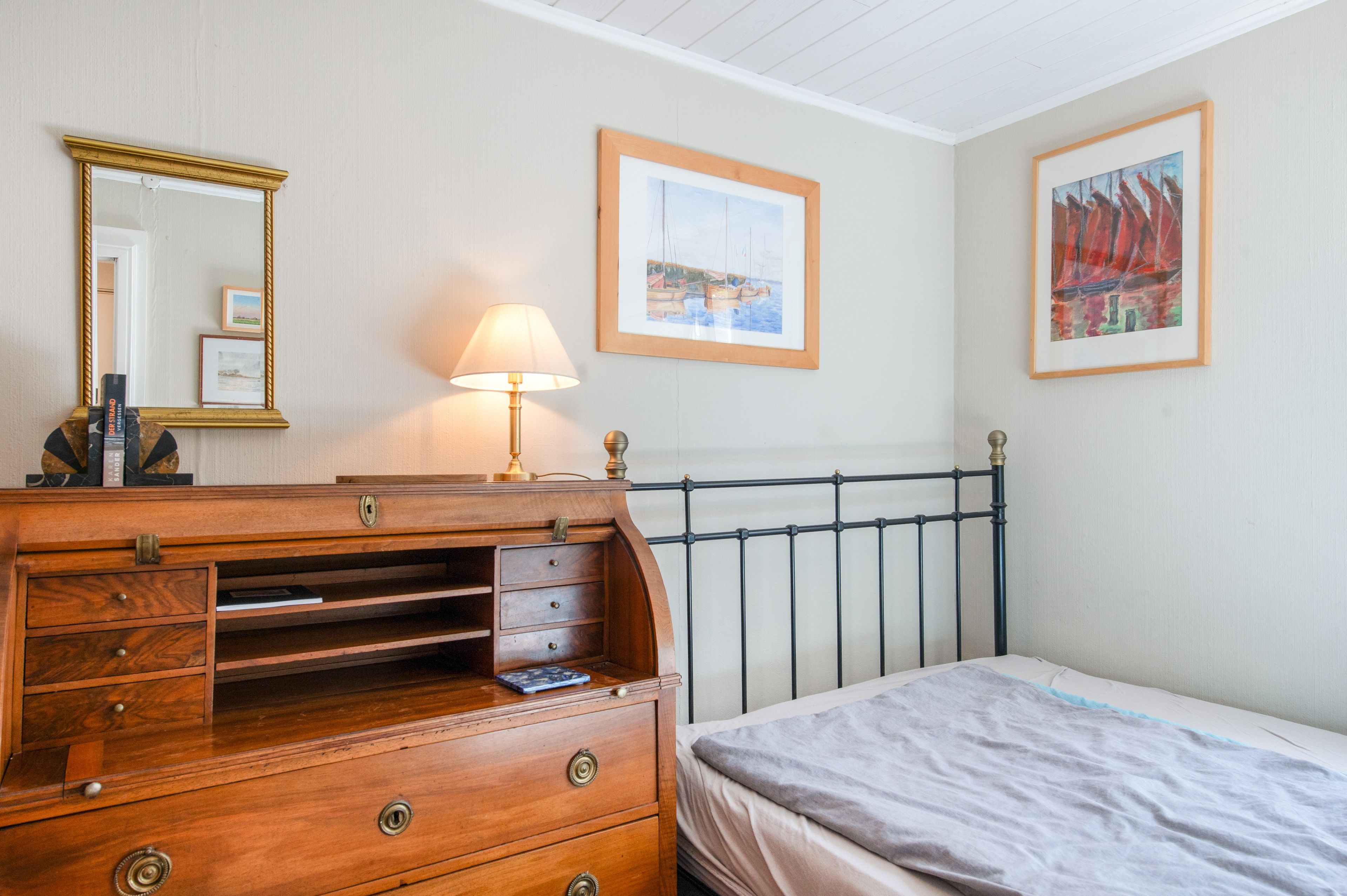 A bedroom with a wooden dresser, a bed, a nightstand, a lamp, a mirror, and two framed pictures on the wall.