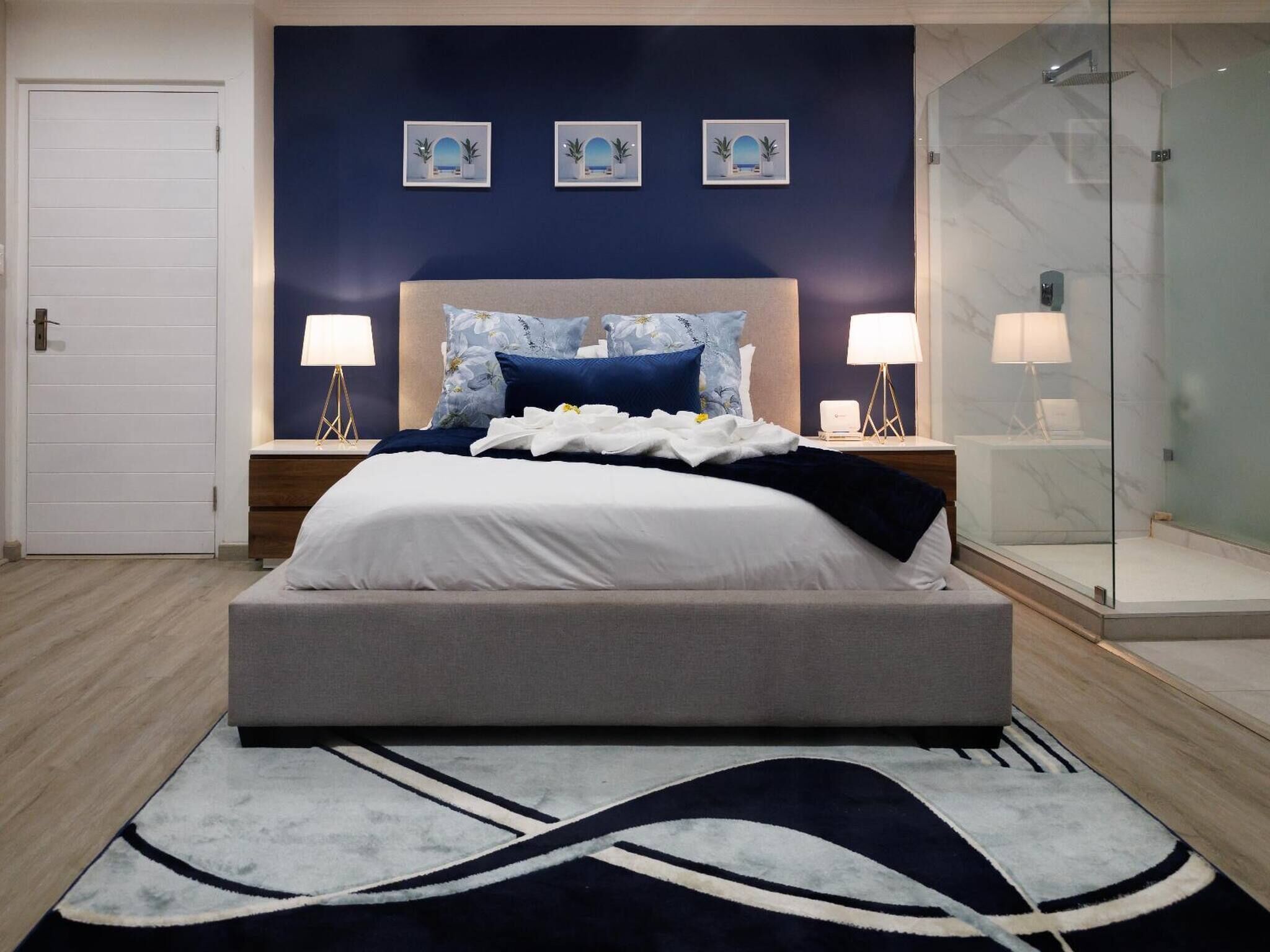 A modern bedroom with a large bed, bedside lamps, a glass shower enclosure, and a view of the sea through framed artwork.