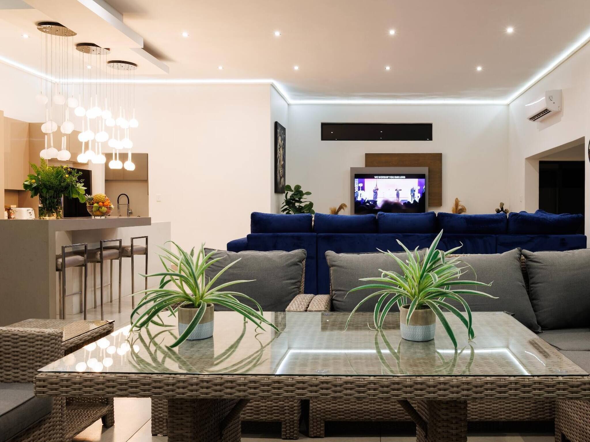 A modern living room with a glass-top dining table, wicker chairs, a blue sofa, and a TV mounted on the wall.