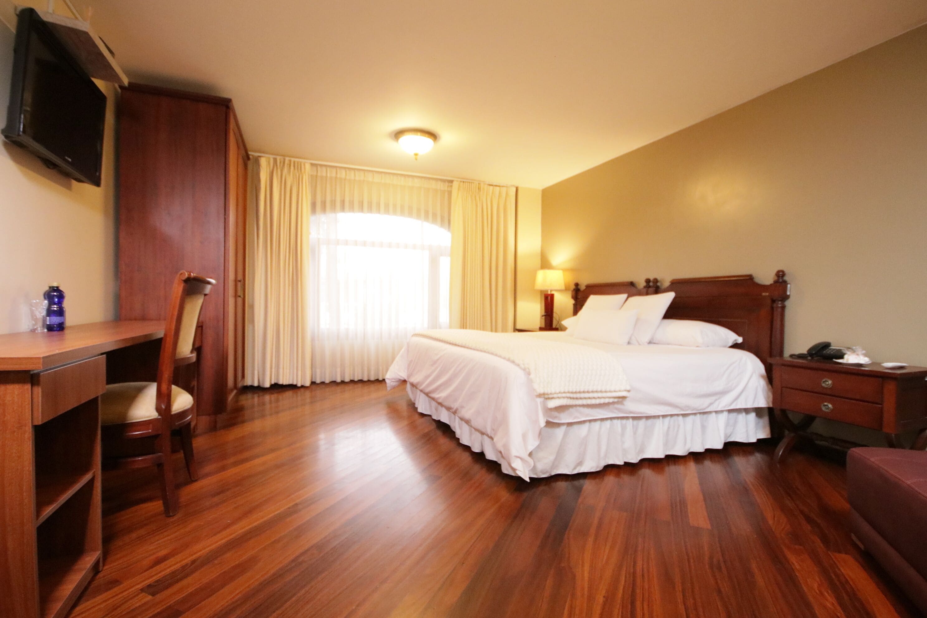 Habitación de hotel con escritorio de madera, cama con ropa blanca, lámpara de noche y televisión montada en la pared.