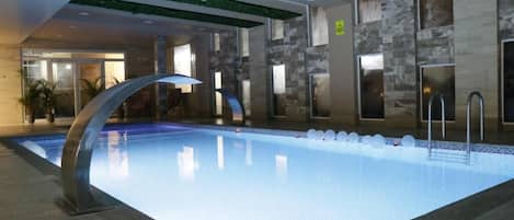Una piscina interior moderna con superficie de agua cristalina, rodeada de paredes de piedra y techo verde.