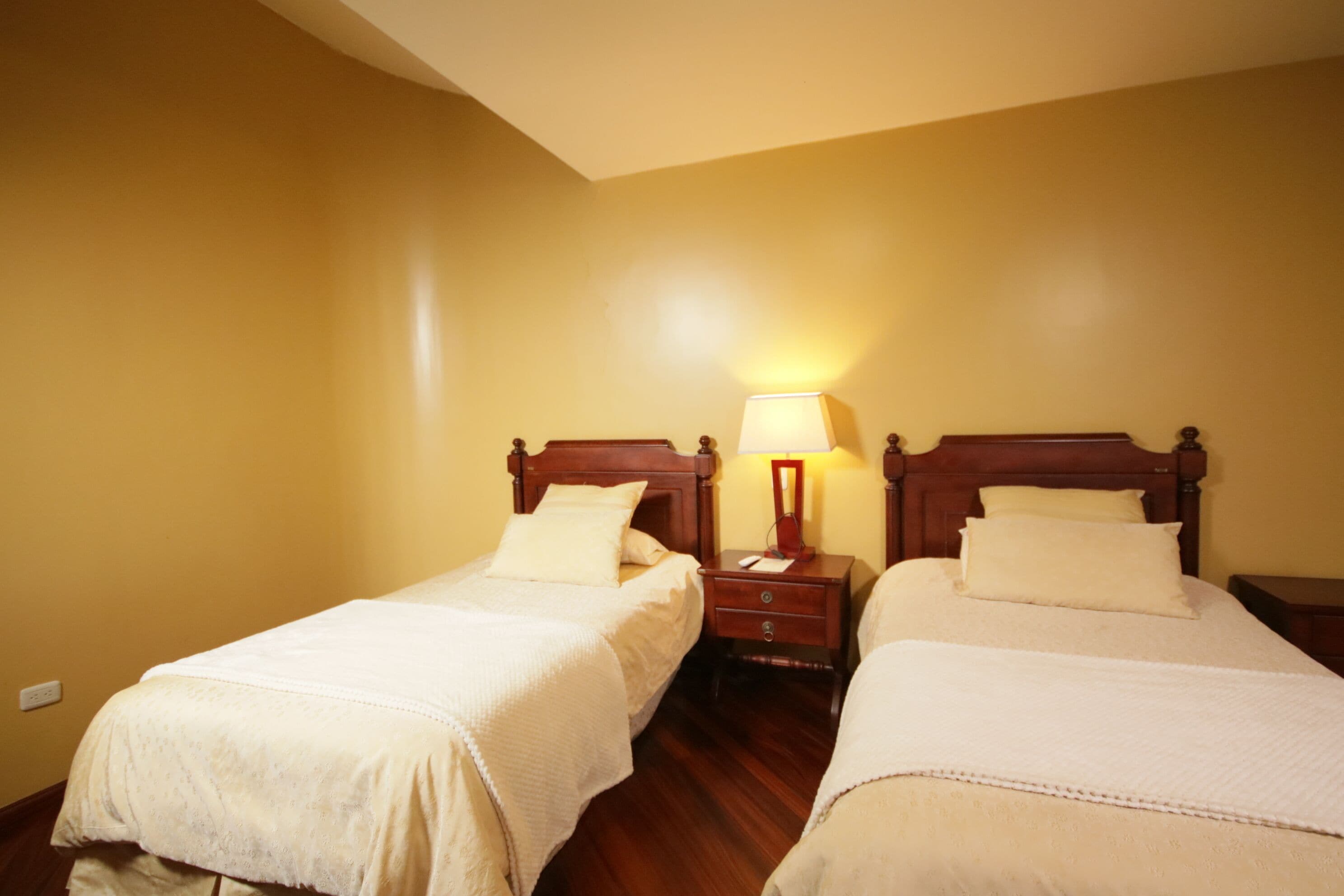 A hotel room with two single beds, wooden headboards, bedside tables with lamps, and plain walls.