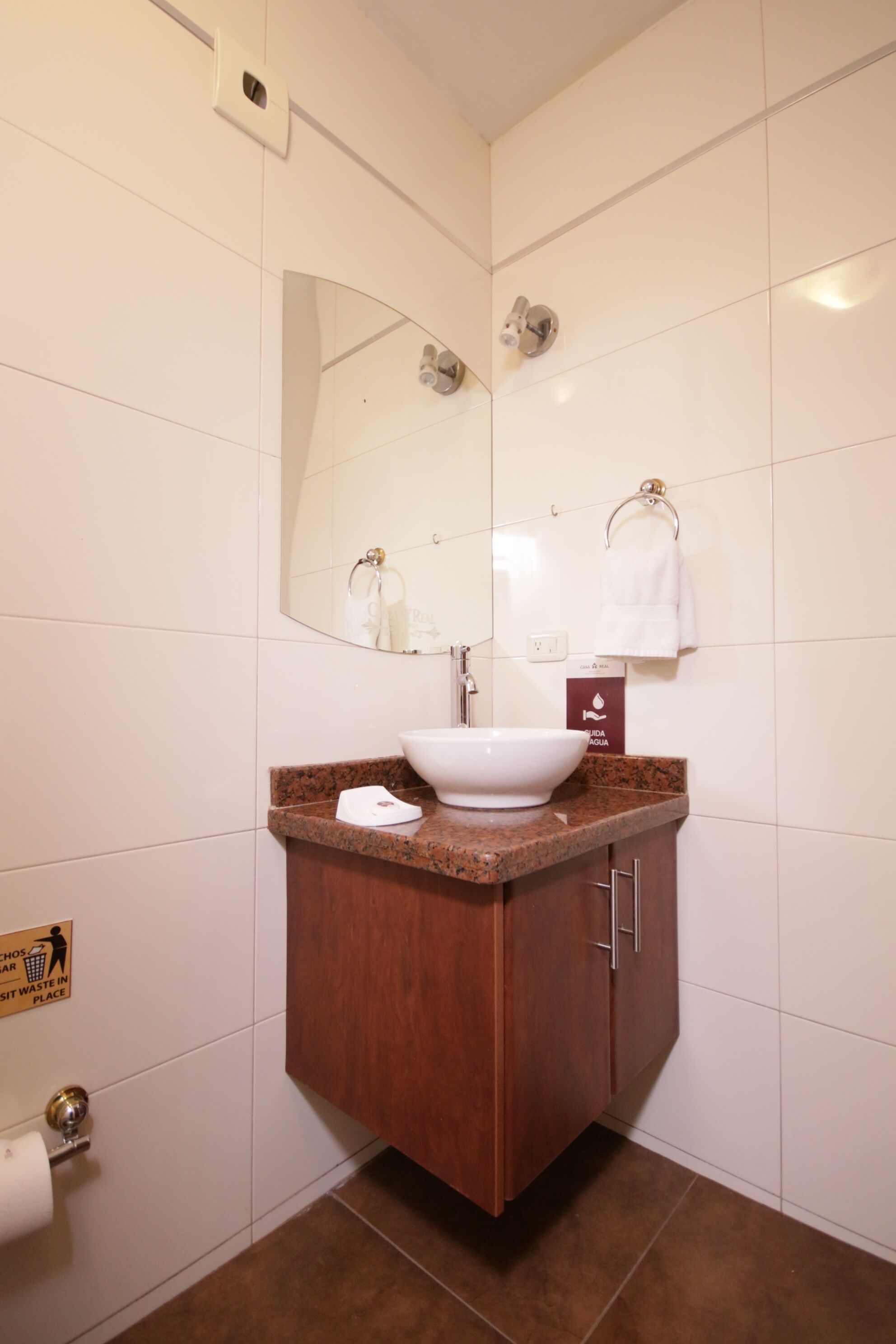A bathroom with a white sink, a mirror, a towel rack, and a sign indicating the location of the trash bin.