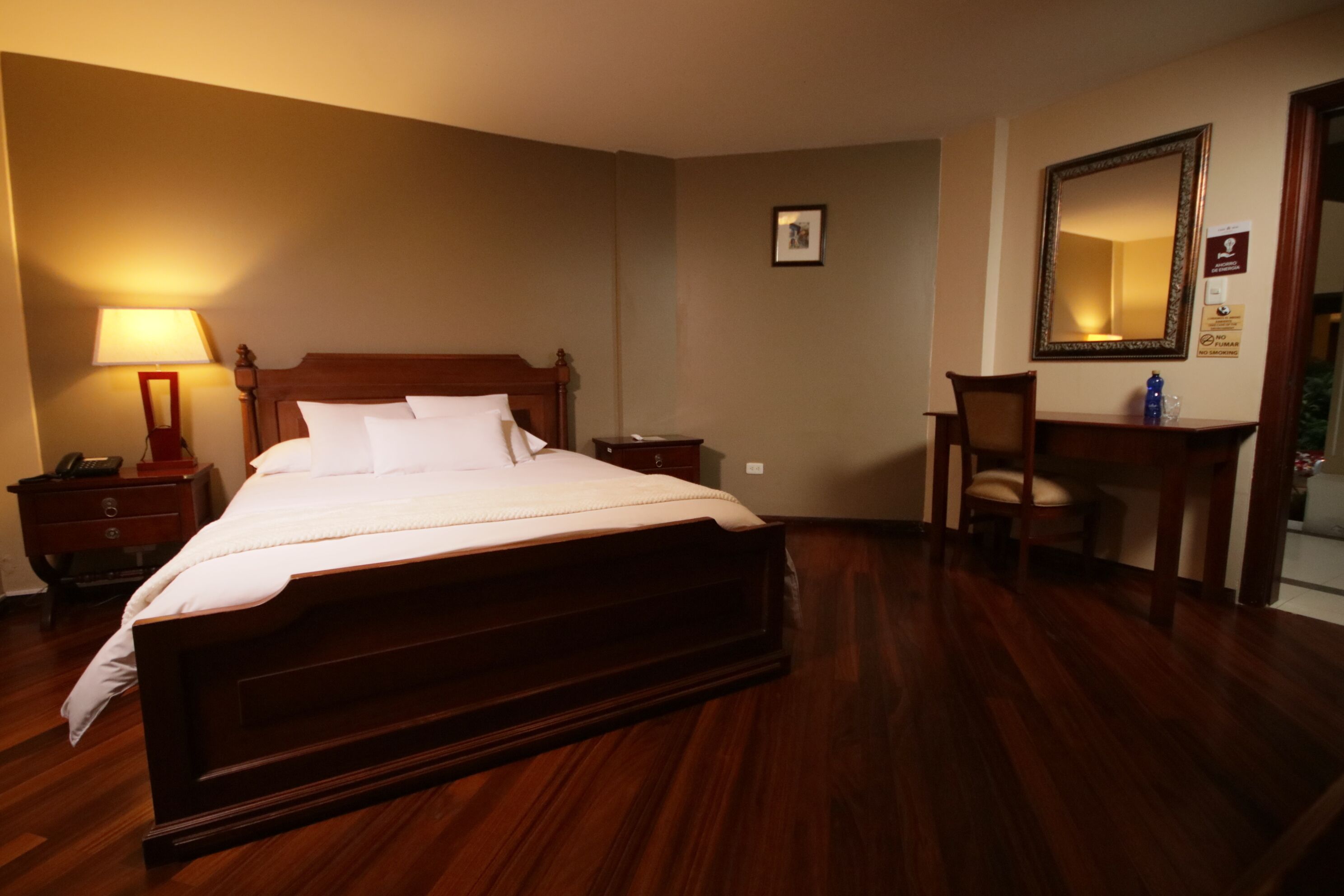 A hotel room with a wooden bed, a desk, a chair, a lamp, and a mirror.