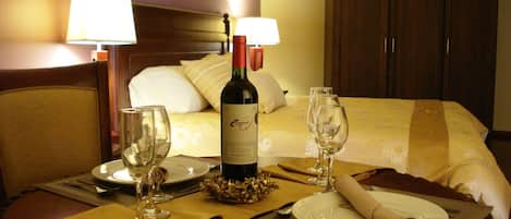 Una mesa de comedor bien puesta con una botella de vino, copas y cubiertos, una cama con almohadas y un armario de madera en una habitación de hotel.