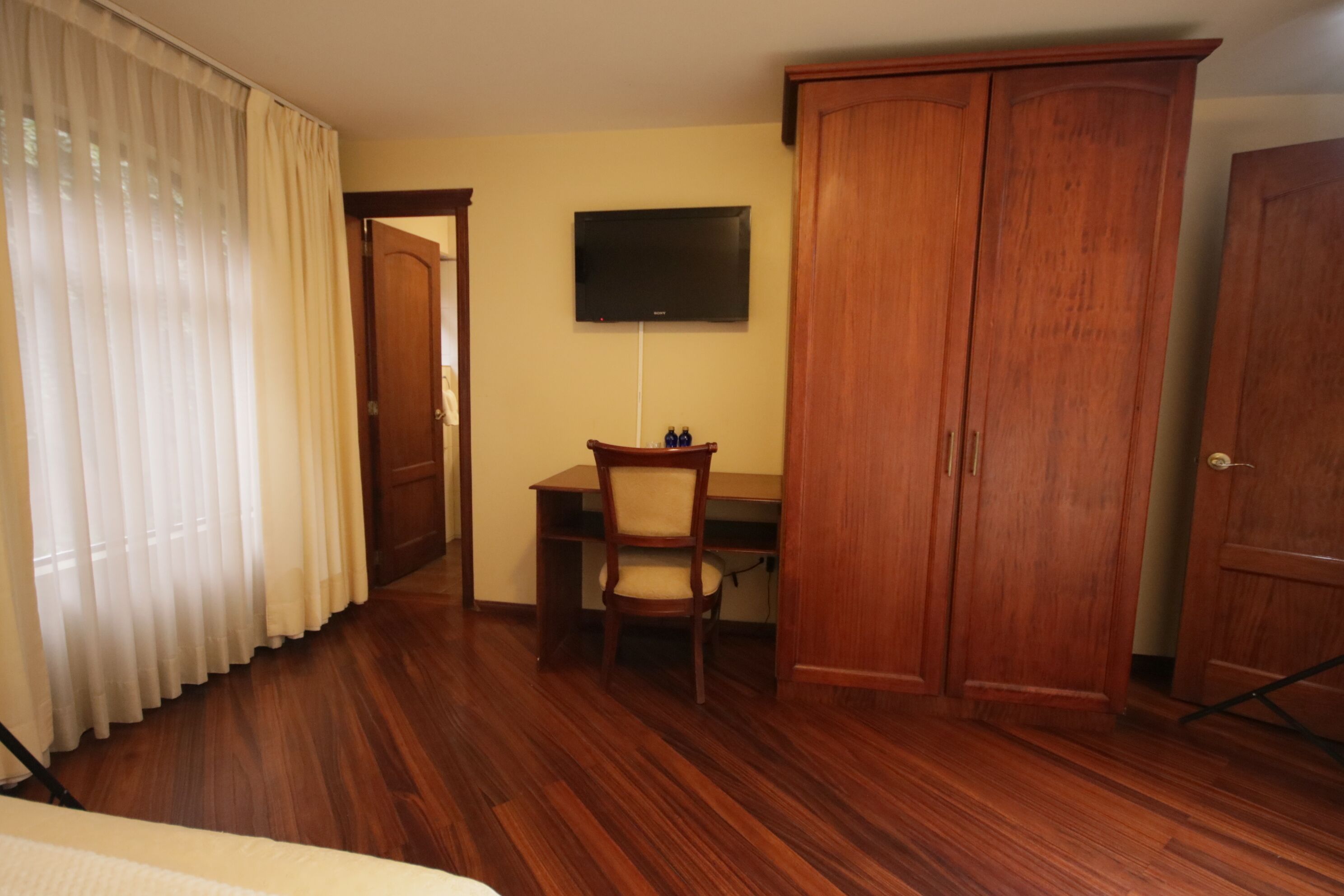 Una habitación con suelo de madera, un escritorio con silla, televisor montado en la pared y un armario.