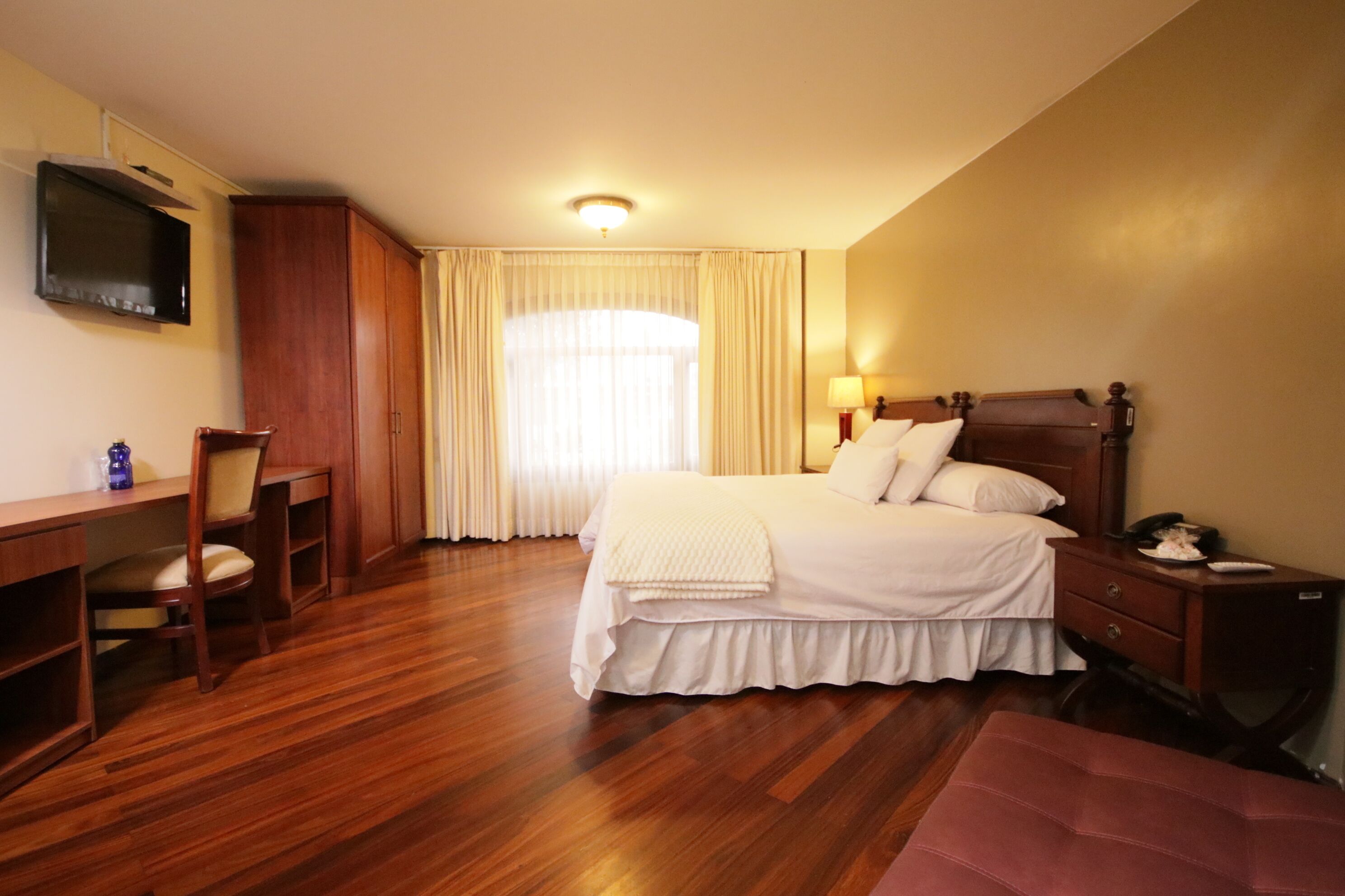 Habitación de hotel con piso de madera, una cama con ropa blanca, un escritorio con una silla, un televisor montado en la pared y una ventana con cortinas.