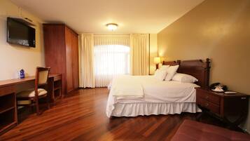 Habitación de hotel con piso de madera, una cama con ropa blanca, un escritorio con una silla, un televisor montado en la pared y una ventana con cortinas.