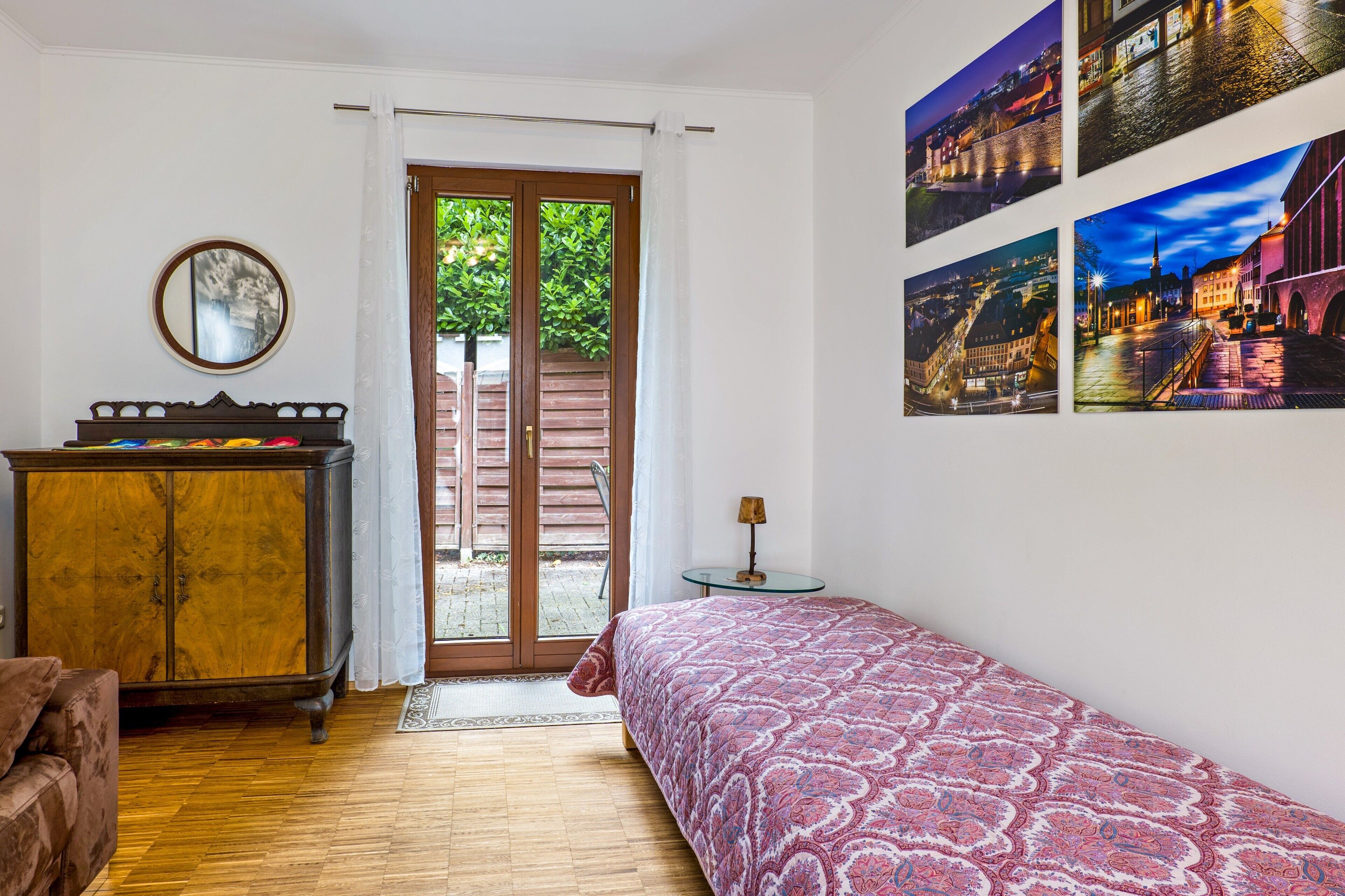 Ein Schlafzimmer mit Holzboden, einem Bett mit gemustertem Überwurf, einem Holzkleiderschrank, einem Spiegel und einer Tür, die zu einem Außenbereich führt.