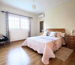 Ein Schlafzimmer mit einem Bett, einem Nachttisch, einem Spiegel, einem Sessel und einem Fenster mit Vorhängen.