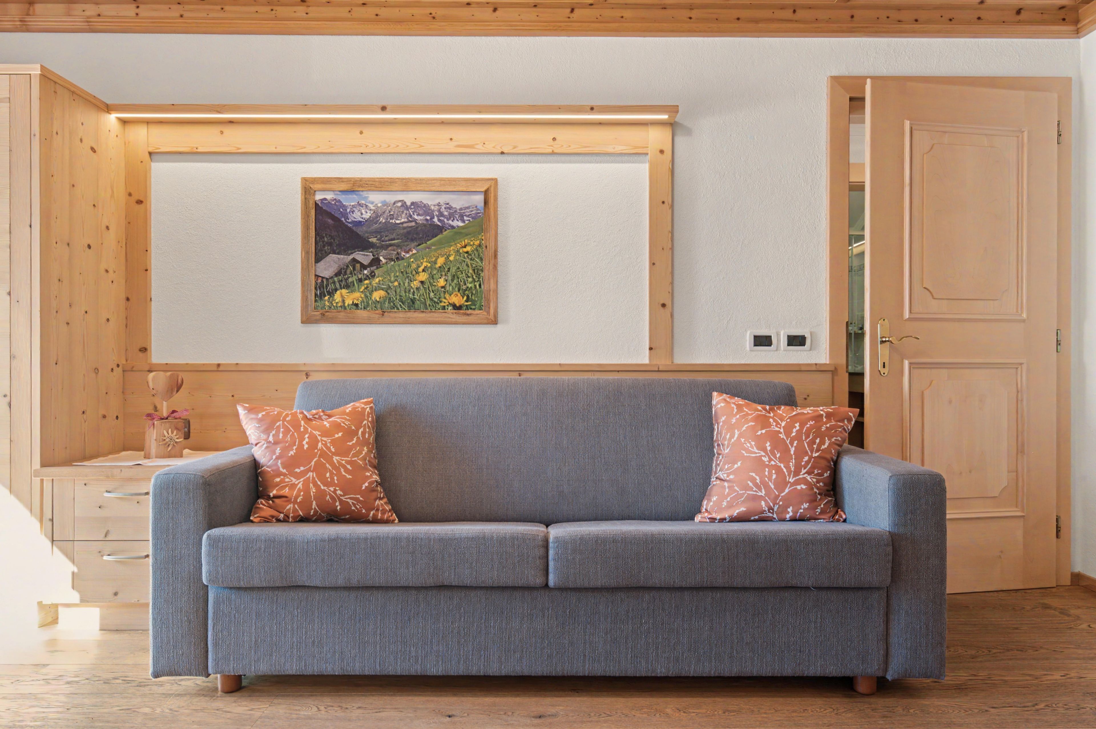 Una stanza con un divano grigio, due cuscini decorati, un quadro di un paesaggio, pannelli in legno e una porta in legno.