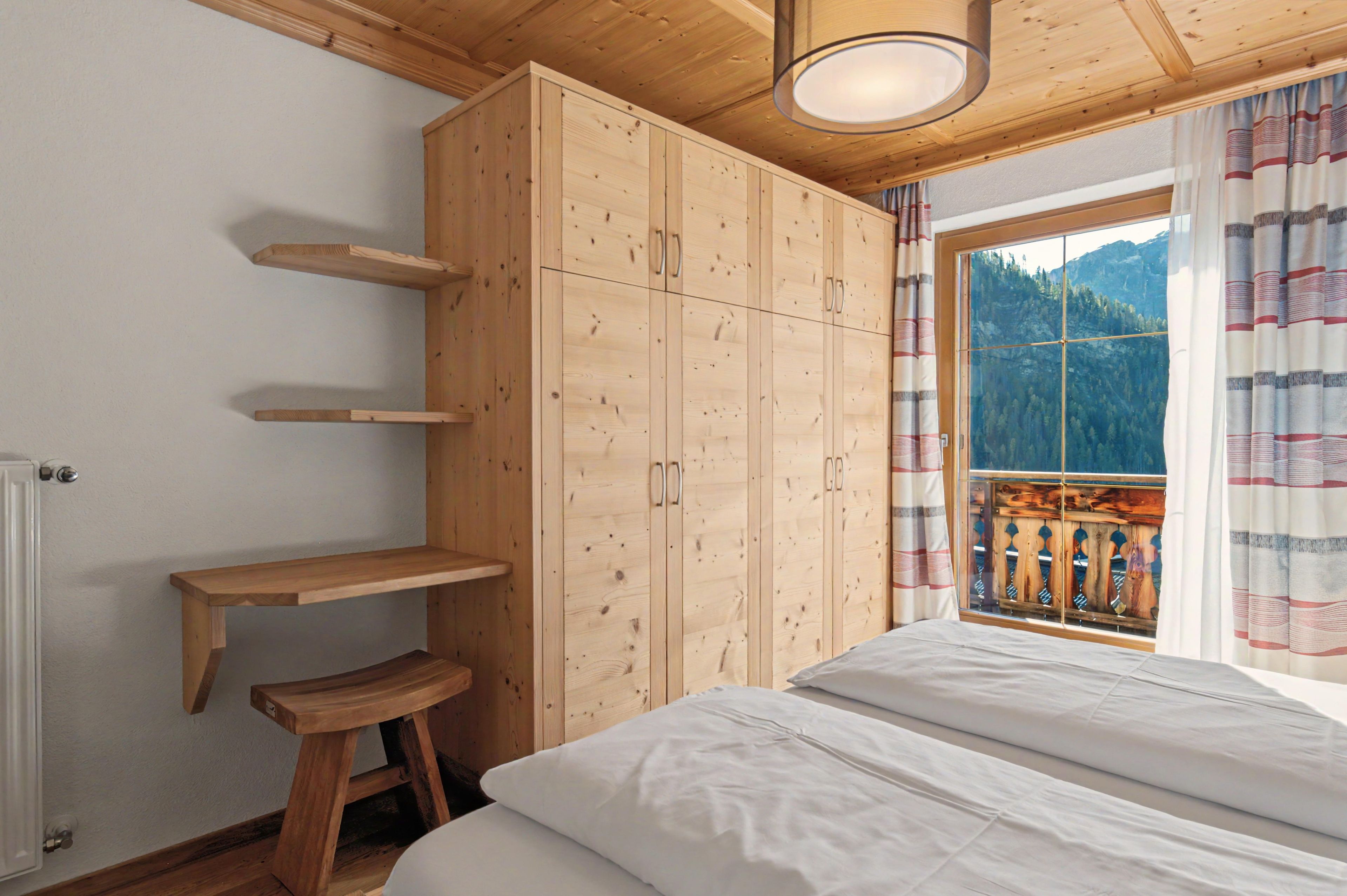 A bedroom with a wooden wardrobe, a bed, a window with a view of mountains, and a wooden stool.