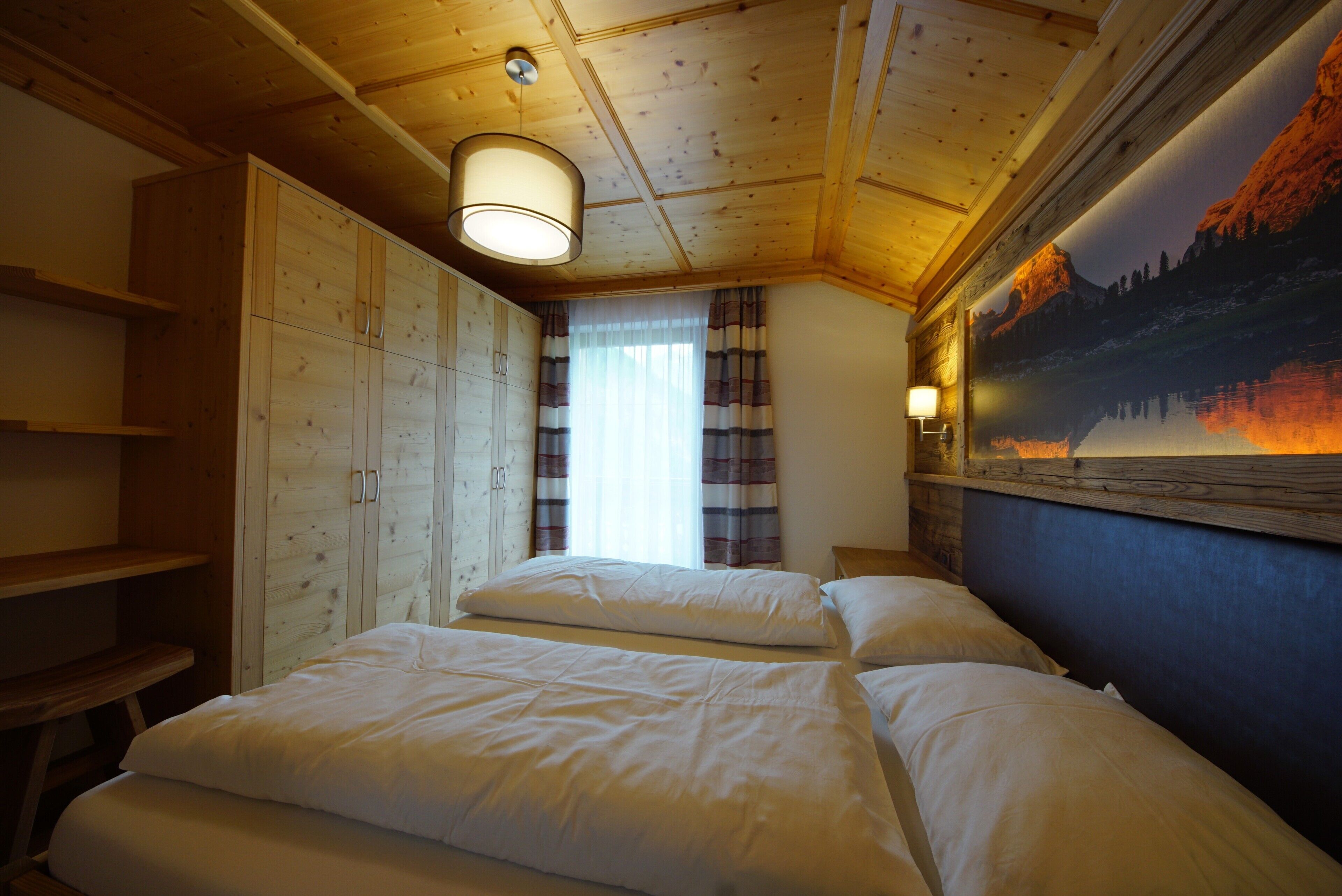 A room with wooden ceiling beams, a bed with white bedding, a window with curtains, and a wall-mounted lamp.