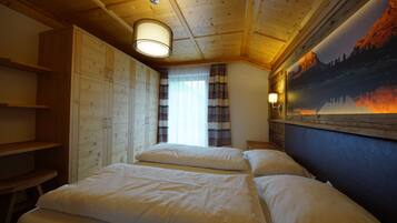 A room with wooden ceiling beams, a bed with white bedding, a window with curtains, and a wall-mounted lamp.