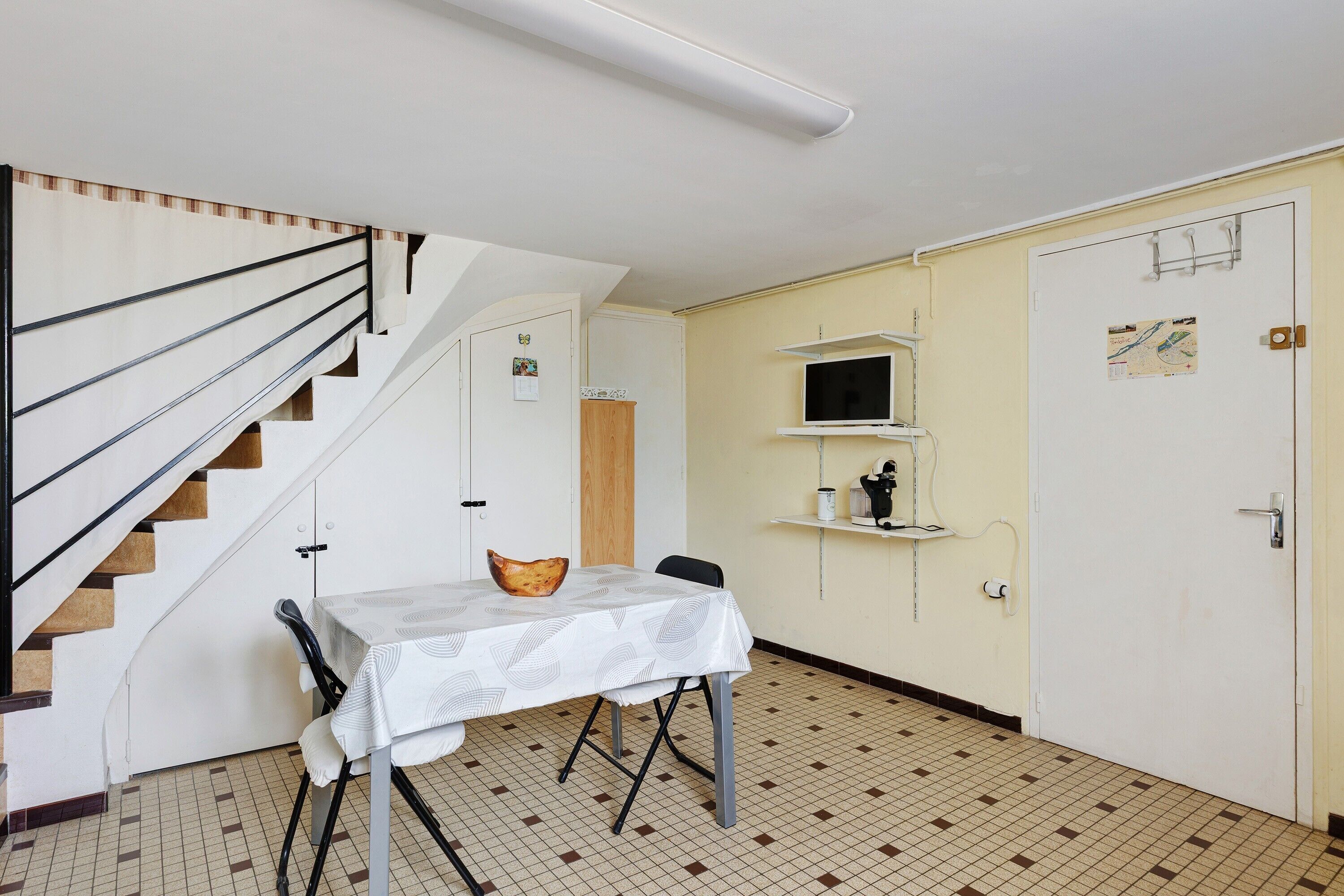 A room with a staircase, a table with a white tablecloth, a chair, a wooden cabinet, and a door with a map.