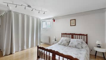 A bedroom with a wooden bed, a window, a radiator, a small table with a lamp, and a curtain.