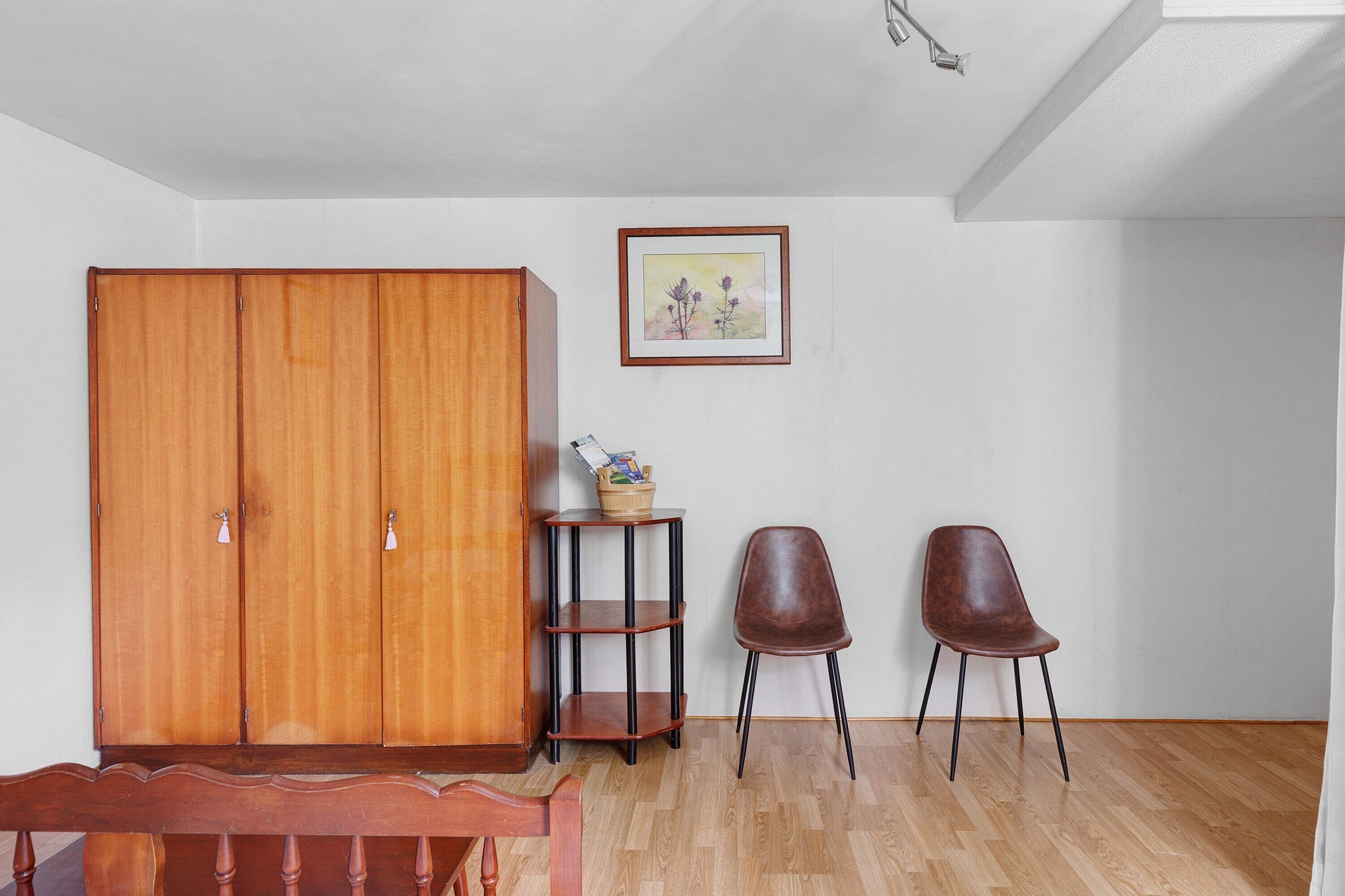 A room with a wooden wardrobe, a small table with a basket, two chairs, and a framed picture on the wall.
