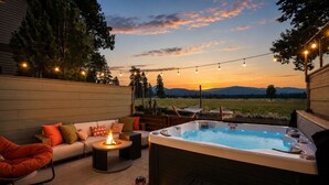 A hot tub with a view of mountains, a sofa with cushions, a fire pit, and string lights.