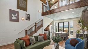 A spacious living area with a green sofa, blue armchair, and a wooden coffee table. A staircase with wooden railings leads to an upper level.
