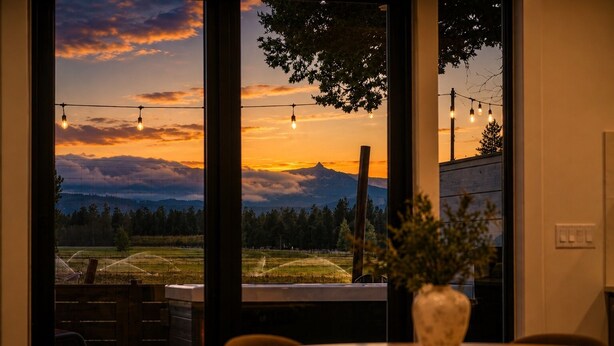 A room with a large window overlooking a landscape at sunset, a table with chairs, and a vase with plants.