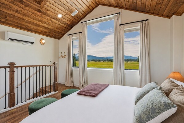 A bedroom with a wooden ceiling, a bed with pillows, a green bench, and a view of a green field and mountains through large windows.