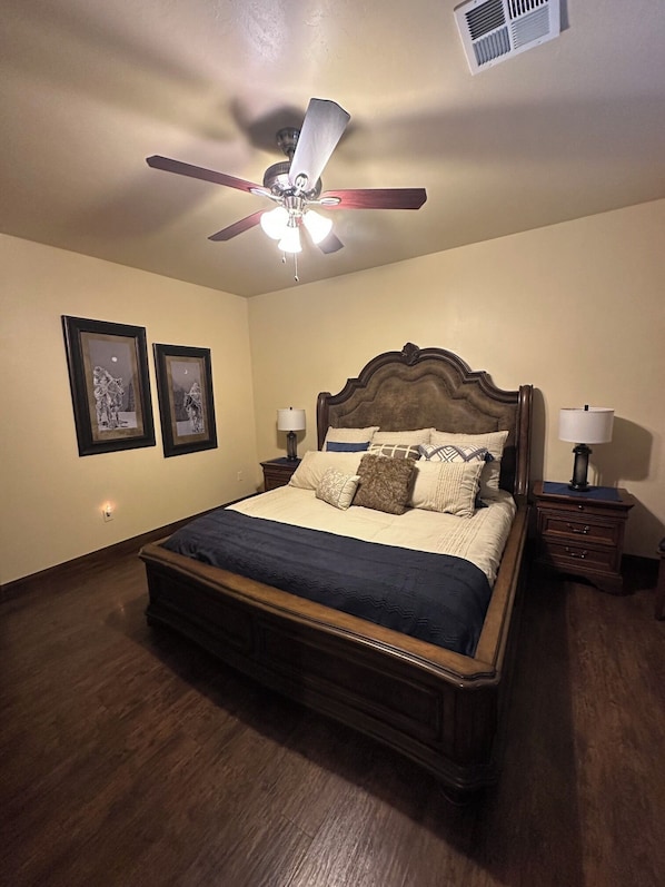 Un dormitorio con una cama de madera, dos mesitas de noche con lámparas, un ventilador de techo y dos cuadros en la pared.