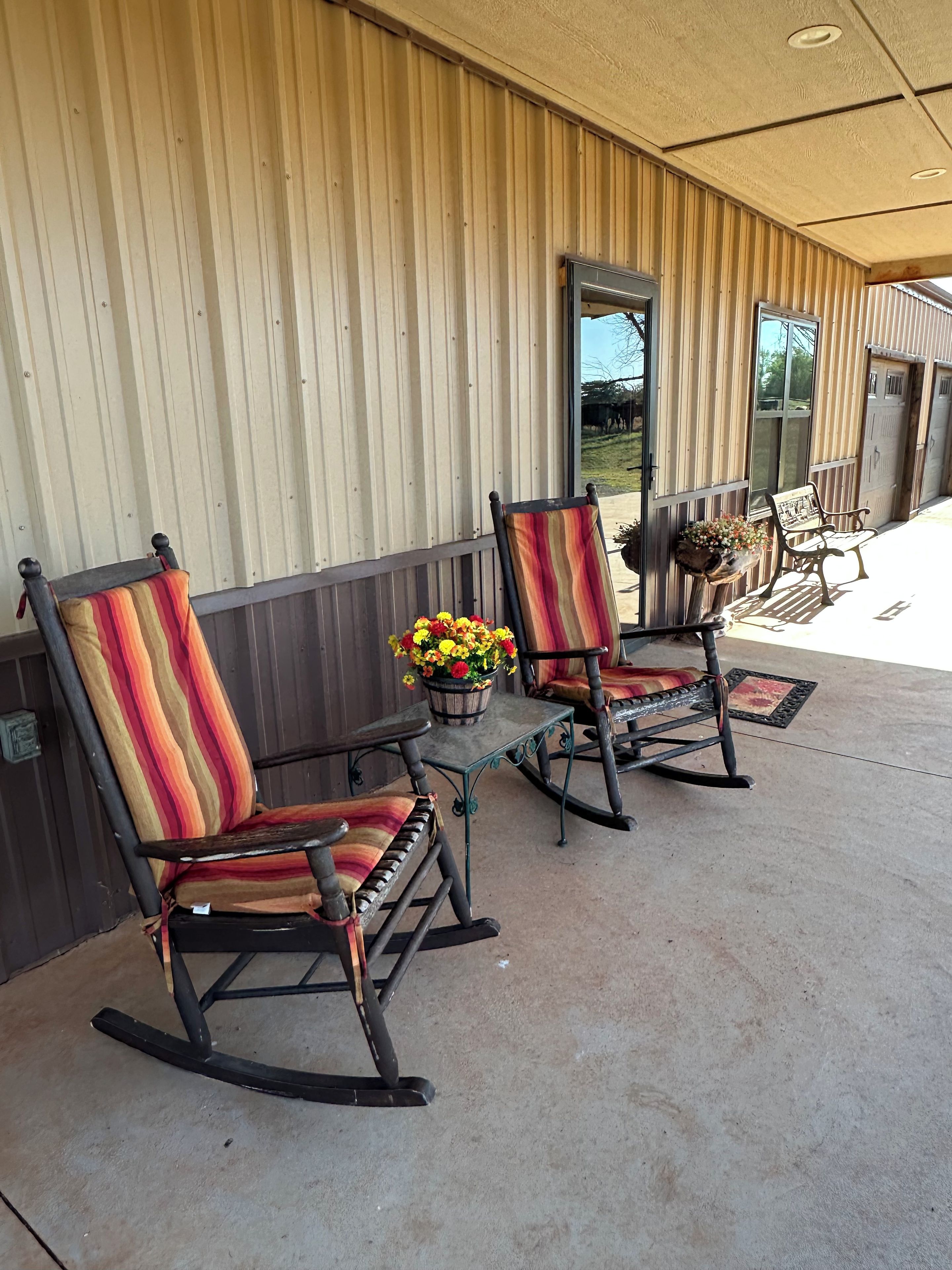 A porch with two rocking chairs and a table, a potted plant, and a wooden building with vertical siding.