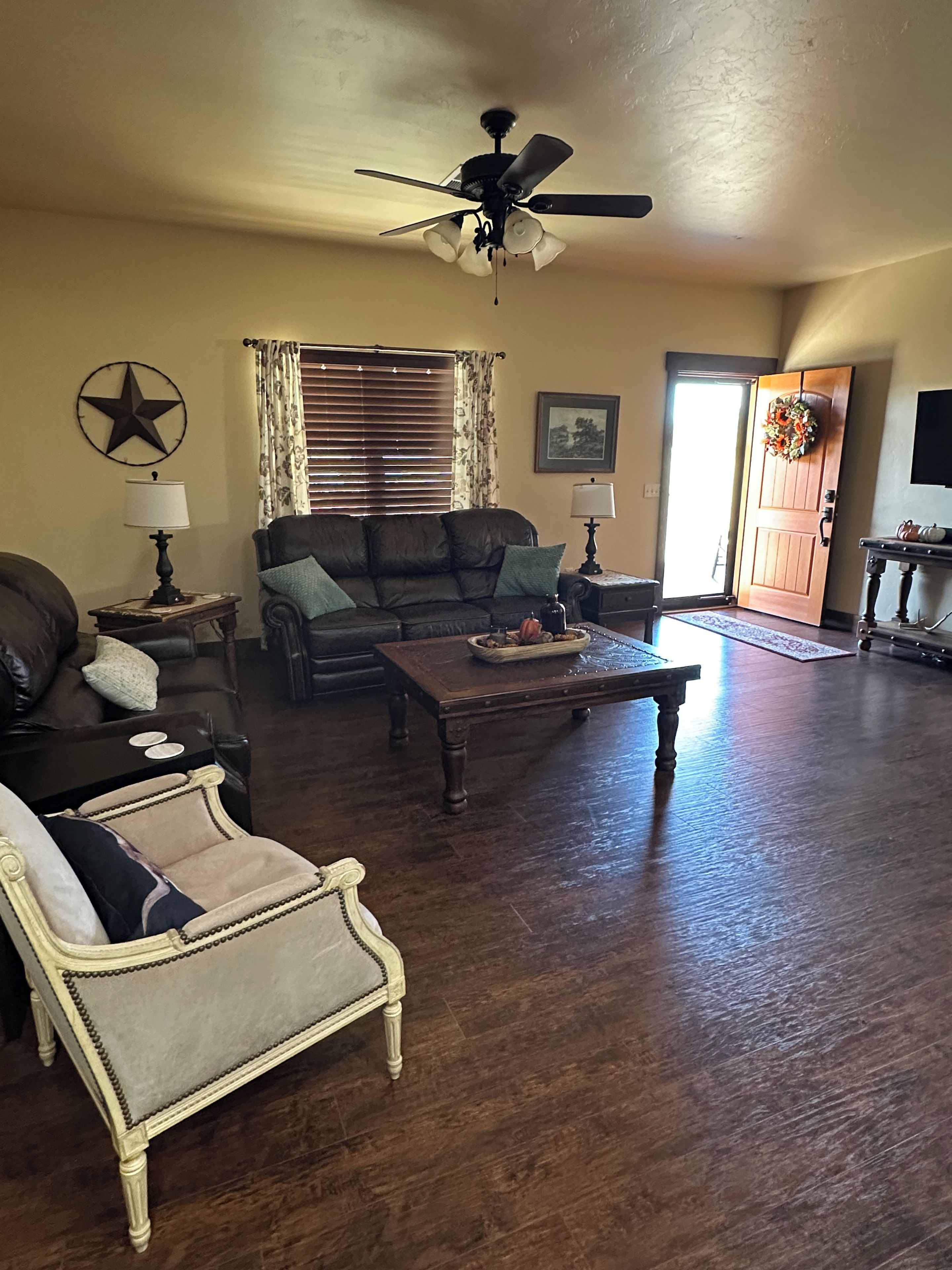 A living room with a ceiling fan, a star decoration, a couch, a chair, a coffee table, and a door with a wreath.