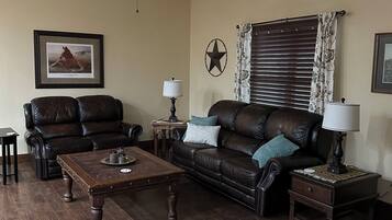 A living room with a brown leather sofa, a wooden coffee table, a ceiling fan, a framed picture on the wall, and a window with blinds.
