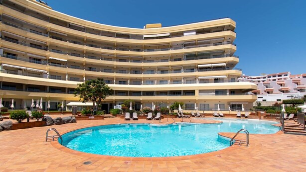Um hotel com vários andares, piscina central rodeada por espreguiçadeiras e guarda-sóis.