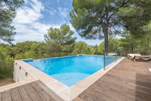 A rectangular swimming pool with a wooden deck and glass railing, surrounded by trees and a clear sky. - Country House 'Bellpuig' with Mountain View, Wi-Fi and Air Conditioning (Artà)