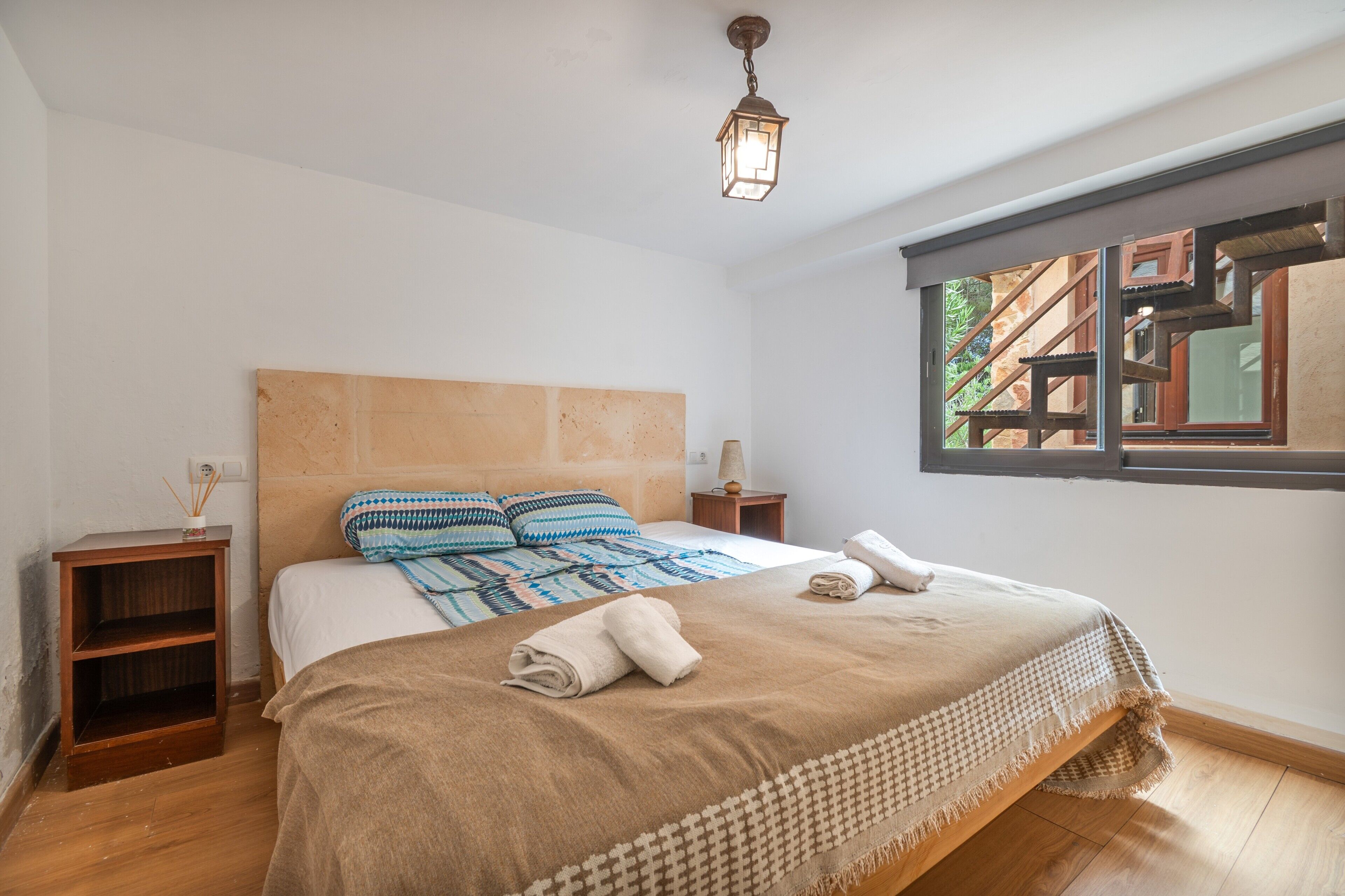 Un dormitorio con una cama grande, un cabecero de madera, una mesita de noche, una lámpara y una ventana con vista a un edificio.