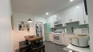 A compact kitchen with white cabinets, a dining table with chairs, a microwave, a stove, and a refrigerator. - Modern 3BR Apartment Near NYC & Cruise Port (Bayonne)