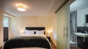 Moderni hotellihuone, jossa on suuri sänky, yöpöydällä lamput, kylpyhuone, jossa on pesuallas ja peili, sekä liukuovi suihkutilaan.