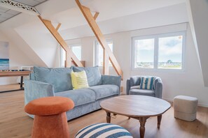 A loft-style room with a high ceiling, exposed wooden beams, a grey sofa, a round wooden table, and a view through large windows. - Villa Josée – Renovated family home, 700 meters from the beach (Kerlouan)