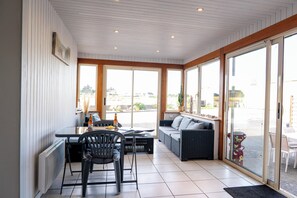 A sunlit room with a dining table, chairs, a sofa, and large windows. - Villa Josée – Renovated family home, 700 meters from the beach (Kerlouan)