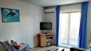 Ein Wohnzimmer mit Fernseher, Sofa, Couchtisch, Sessel und einem Fenster mit Vorhängen.