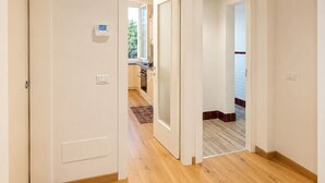 A hallway with wooden flooring, white walls, and a view into a kitchen area with wooden cabinets and a window. - Modern Terrace Retreat (Florence)