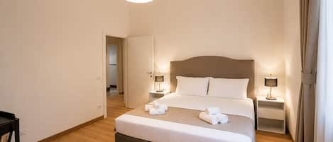 A hotel room with a bed, bedside tables, a hanging light fixture, and a door leading to another room.