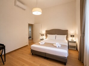 Ett hotellrum med en säng, sängbord, en hängande lampa och en dörr som leder till ett annat rum.