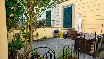 Un patio con un árbol pequeño, plantas en macetas, una mesa con sillas y un edificio amarillo con contraventanas verdes.