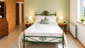 Ein Schlafzimmer mit grünem Metallbettgestell, weißer Bettwäsche, zwei Nachttischlampen, einer Holzkommode und Blick ins Grüne durch ein Fenster.