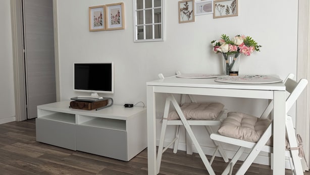 A modern room with a white TV stand, a white dining table with chairs, a white arched window, and framed botanical prints on the wall.