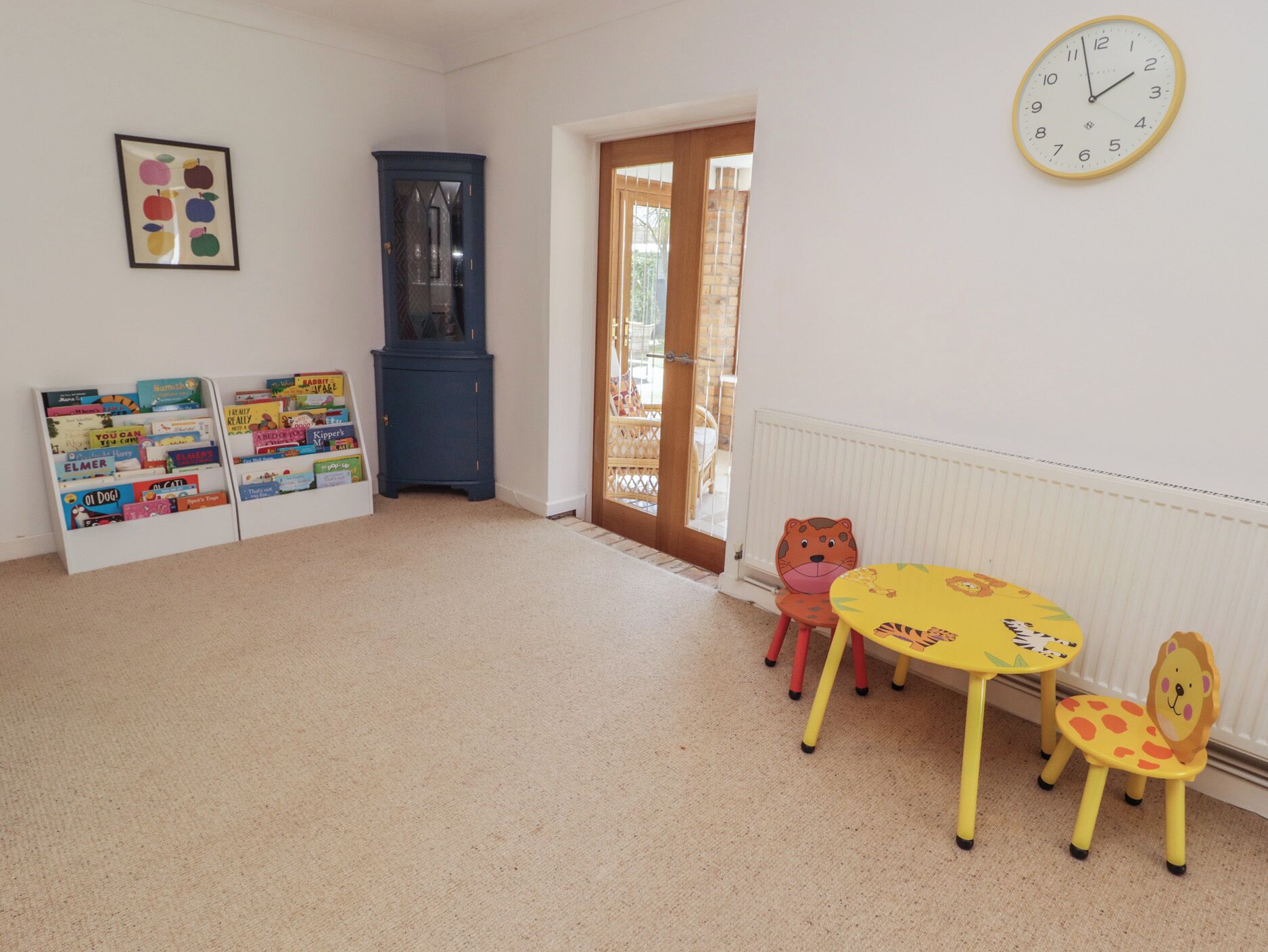 Une pièce avec un meuble en bois, une bibliothèque remplie de livres pour enfants, une table ronde avec des chaises et une horloge murale.