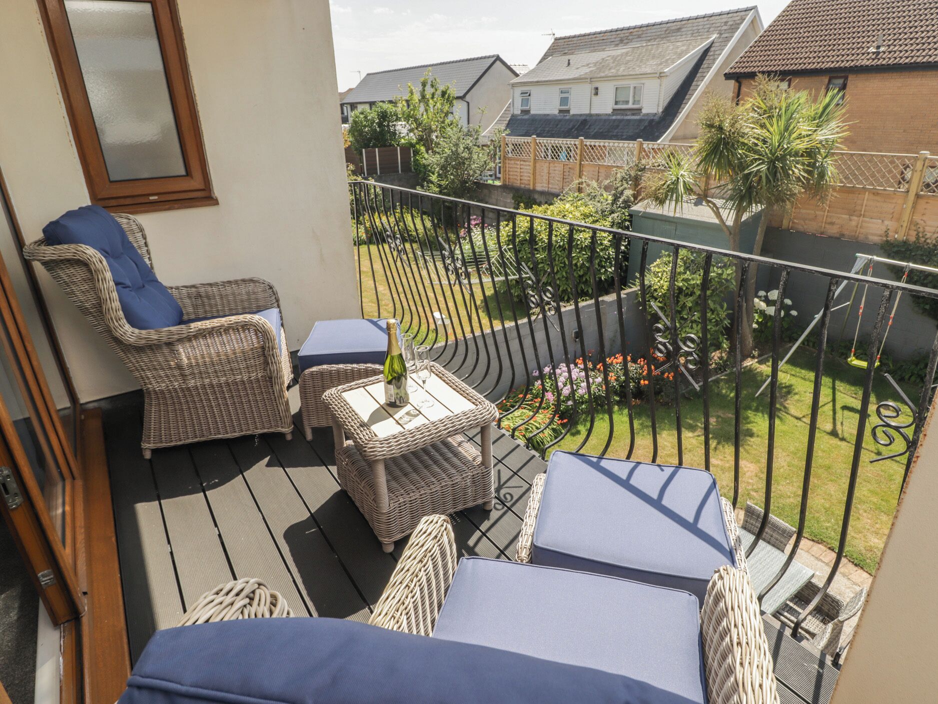 A balcony with wicker furniture, a small table with a bottle, and a view of a garden with a fence.